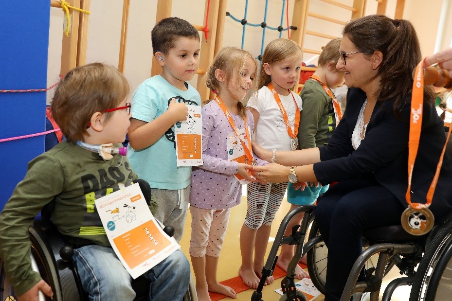 Kinder bekommen von einer jungen Frau im Rollstuhl Mini-Sportabzeichen-Urkunden und Medaillen überreicht. 